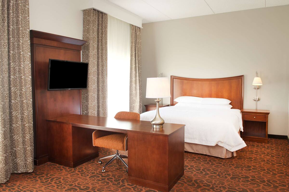 Une chambre d’hôtel avec un bureau en bois, une télévision, un lit avec du linge de maison blanc, une lampe et une moquette à motifs.