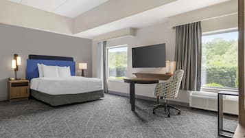 A hotel room with a bed, a desk, a chair, a TV, and a window with a view of greenery.