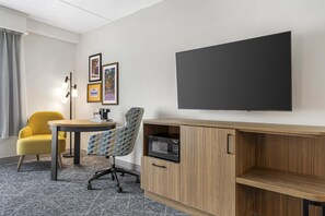 A hotel room with a flat-screen TV, a wooden desk, a yellow armchair, a patterned armchair, a coffee machine, and framed pictures on the wall.