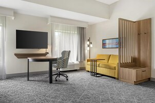 Habitación de hotel con televisión de pantalla plana, sofá, una silla, una mesita pequeña y un armario de madera.