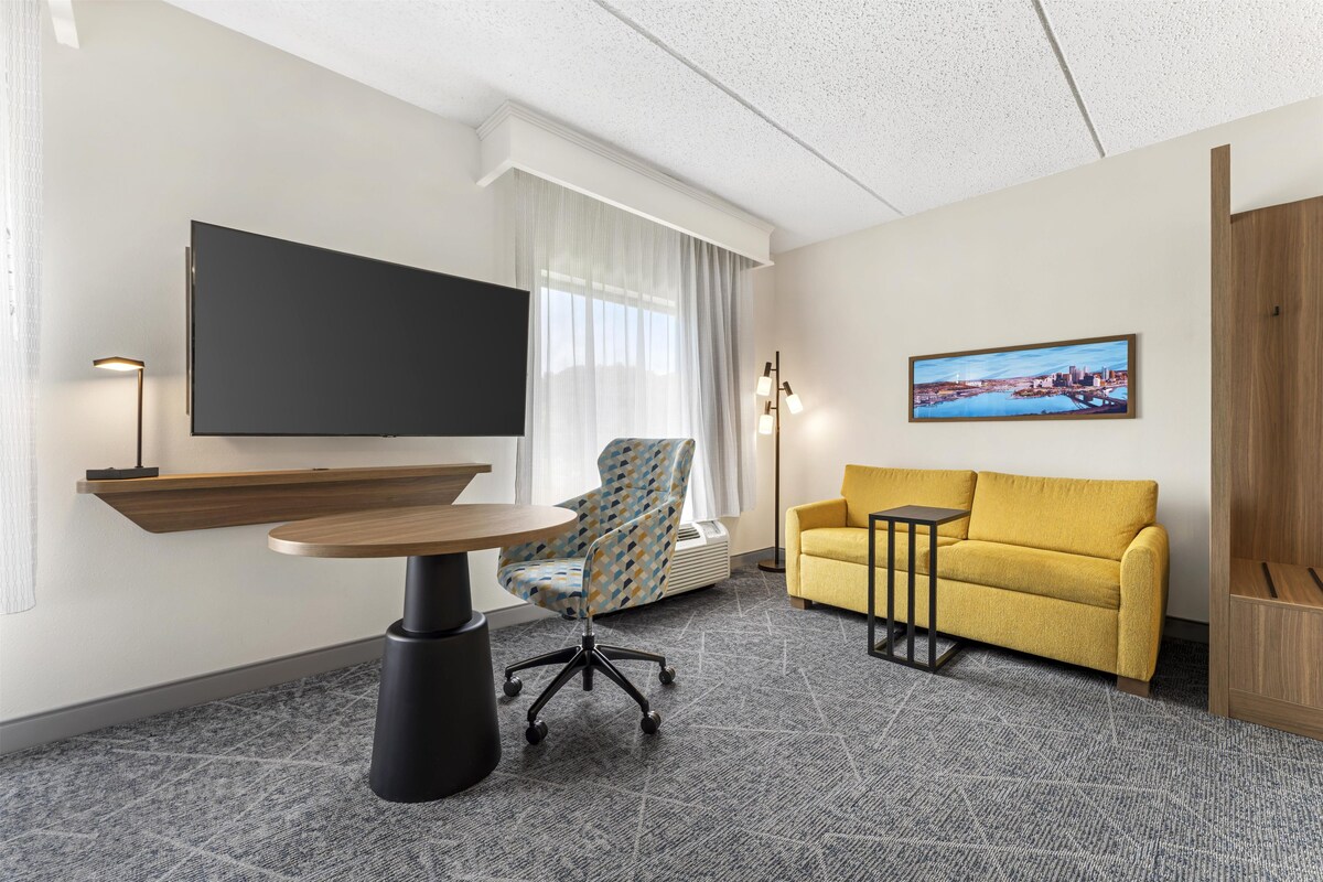 A hotel room with a flat-screen TV, a sofa, a small table, and a chair.