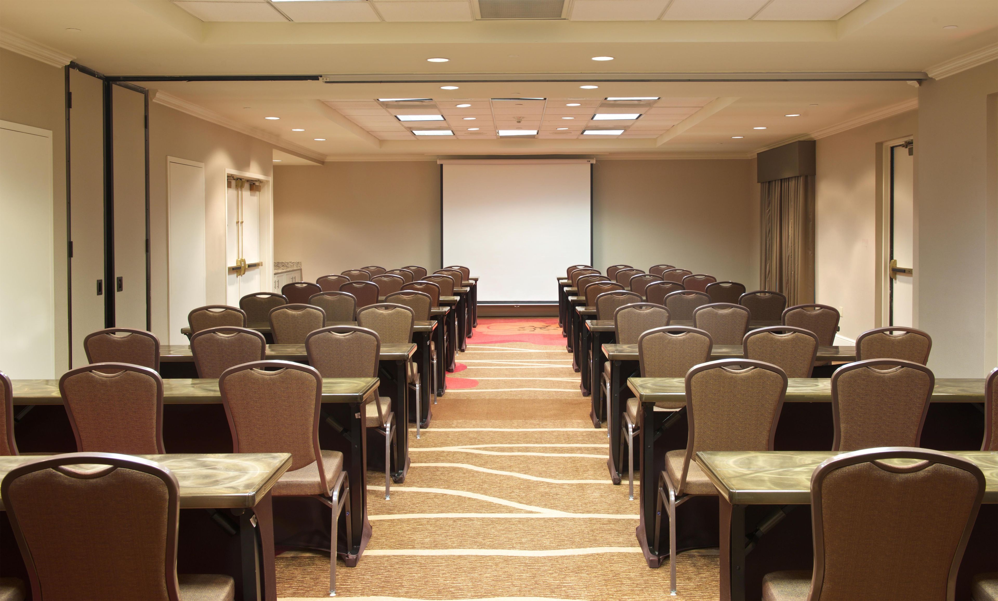 A conference room with rows of tables and chairs, a projector screen, and a carpeted floor.