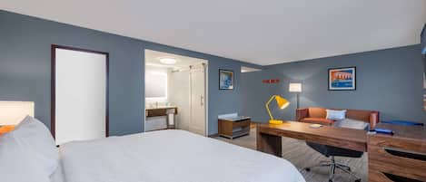 A hotel room with a large bed, a desk with a chair, a desk lamp, a nightstand, and a bathroom visible through an open door.