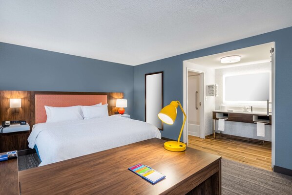 A hotel room with a bed, bedside lamps, a desk with a lamp, a bathroom with a sink and mirror, and a door leading to another room.
