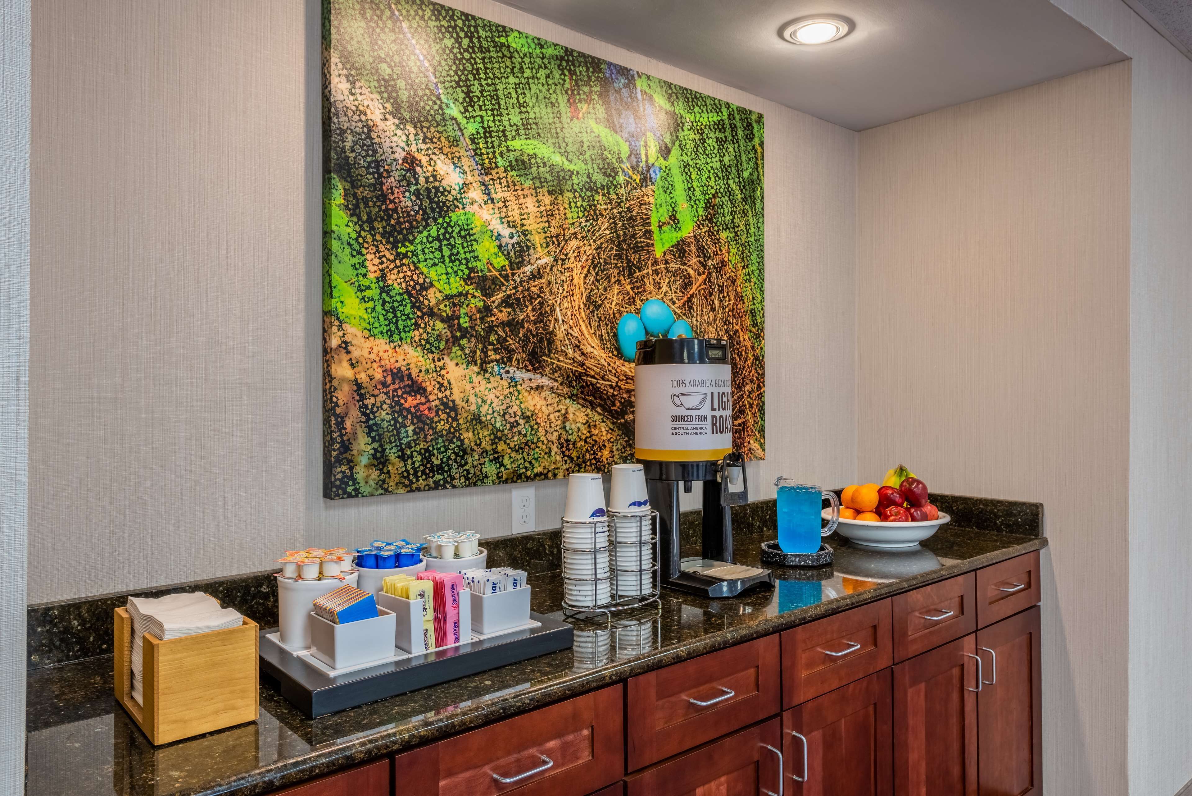 Une cuisine d’hôtel équipée d’un coin café, d’un panier de fruits et d’un grand tableau abstrait accroché au mur.
