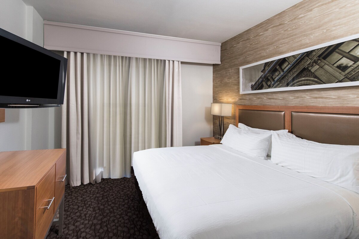 A hotel room with a bed, a TV, a wooden dresser, a nightstand, a lamp, and a painting on the wall.