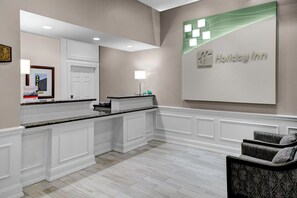 A hotel lobby with a reception desk, a large Holiday Inn sign, a framed picture on the wall, and a seating area with two chairs. - Holiday Inn Savannah Historic District by IHG (Savannah)
