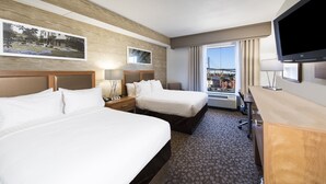 Ein Hotelzimmer mit zwei Betten, einem Schreibtisch, einem Fernseher und einem Fenster mit Blick auf die Stadt.