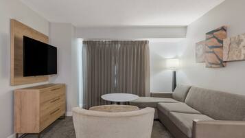 A modern hotel room with a flat-screen TV, wooden dresser, beige sofa, round table, and a large window with curtains.