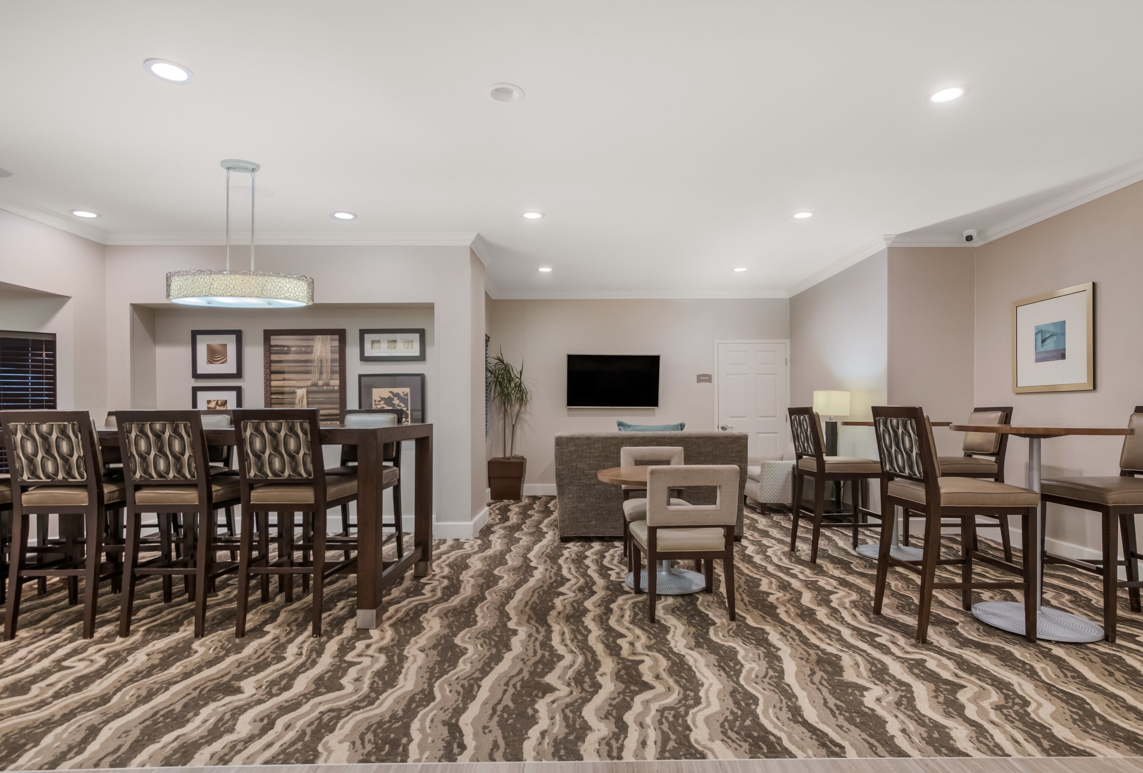 A spacious living room with a patterned carpet, a flat-screen TV mounted on the wall, and a dining area with chairs and a table.