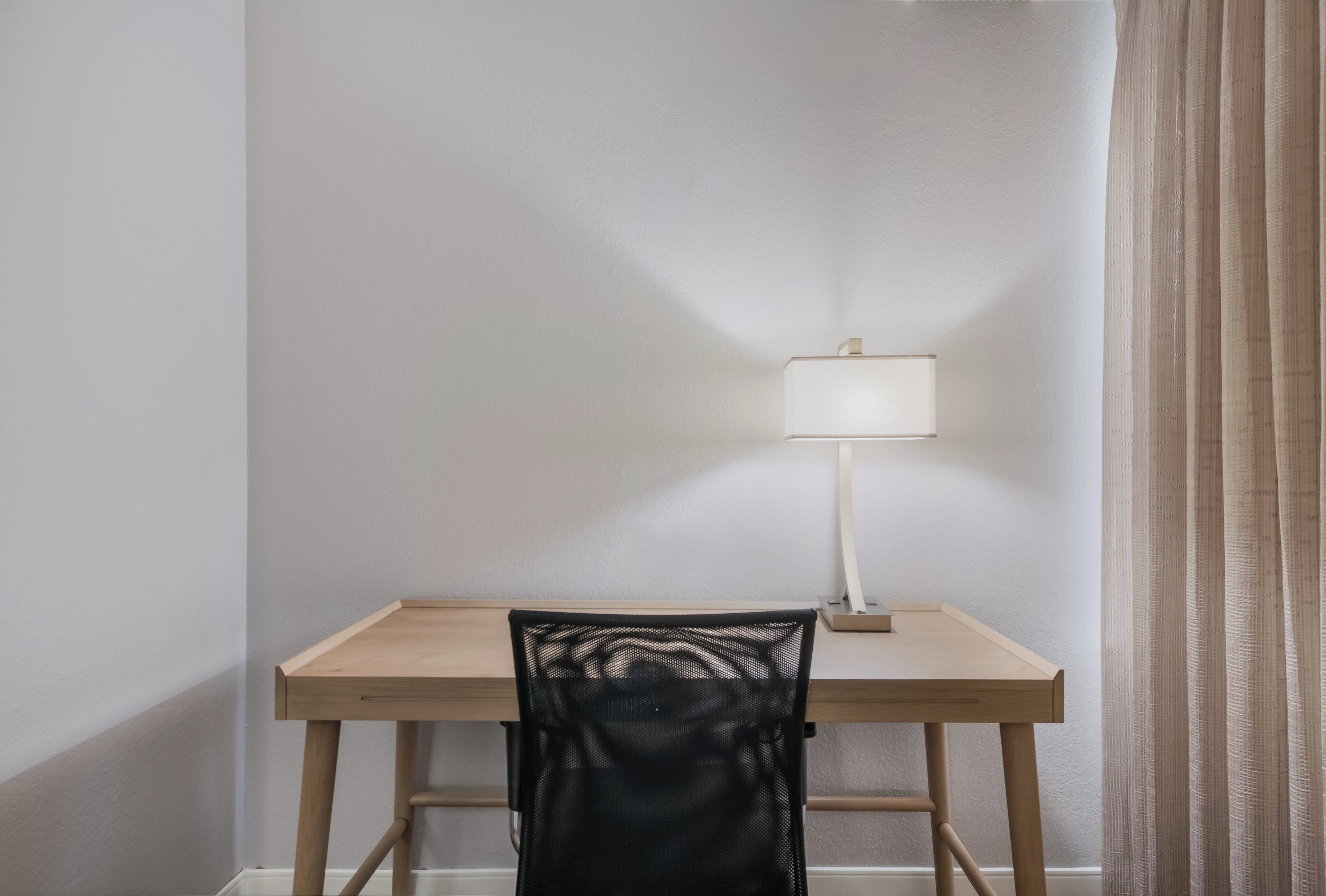 Un escritorio sencillo con una silla y una lámpara en una habitación con paredes lisas y un ventanal con cortinas.