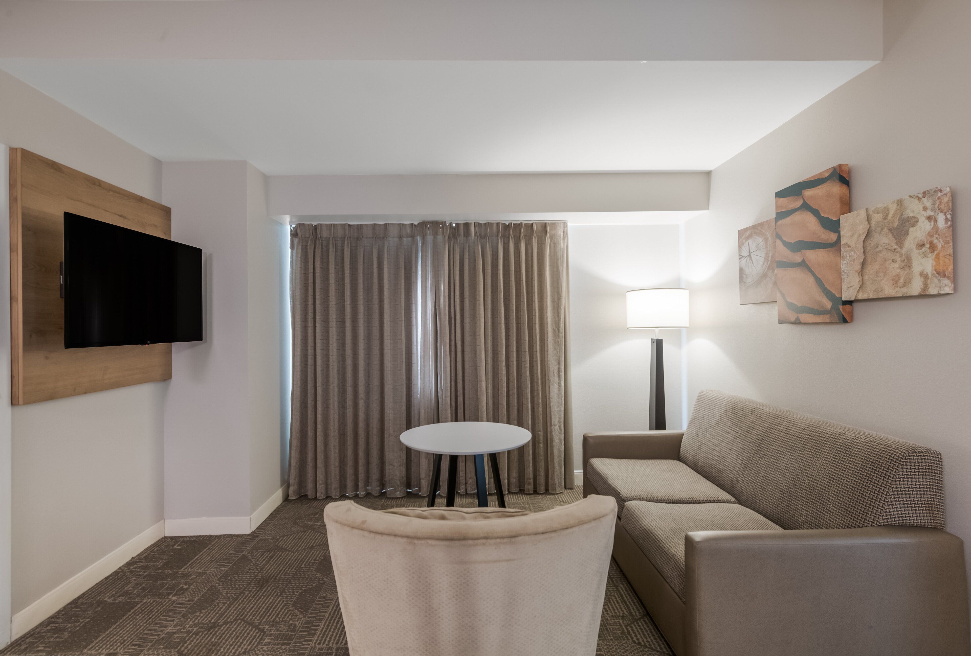 Una habitación de hotel moderna con una televisión de pantalla plana montada en la pared, un sofá, una pequeña mesa redonda y un amplio ventanal con cortinas.