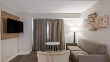 Una habitación de hotel moderna con una televisión de pantalla plana montada en la pared, un sofá, una pequeña mesa redonda y un amplio ventanal con cortinas.