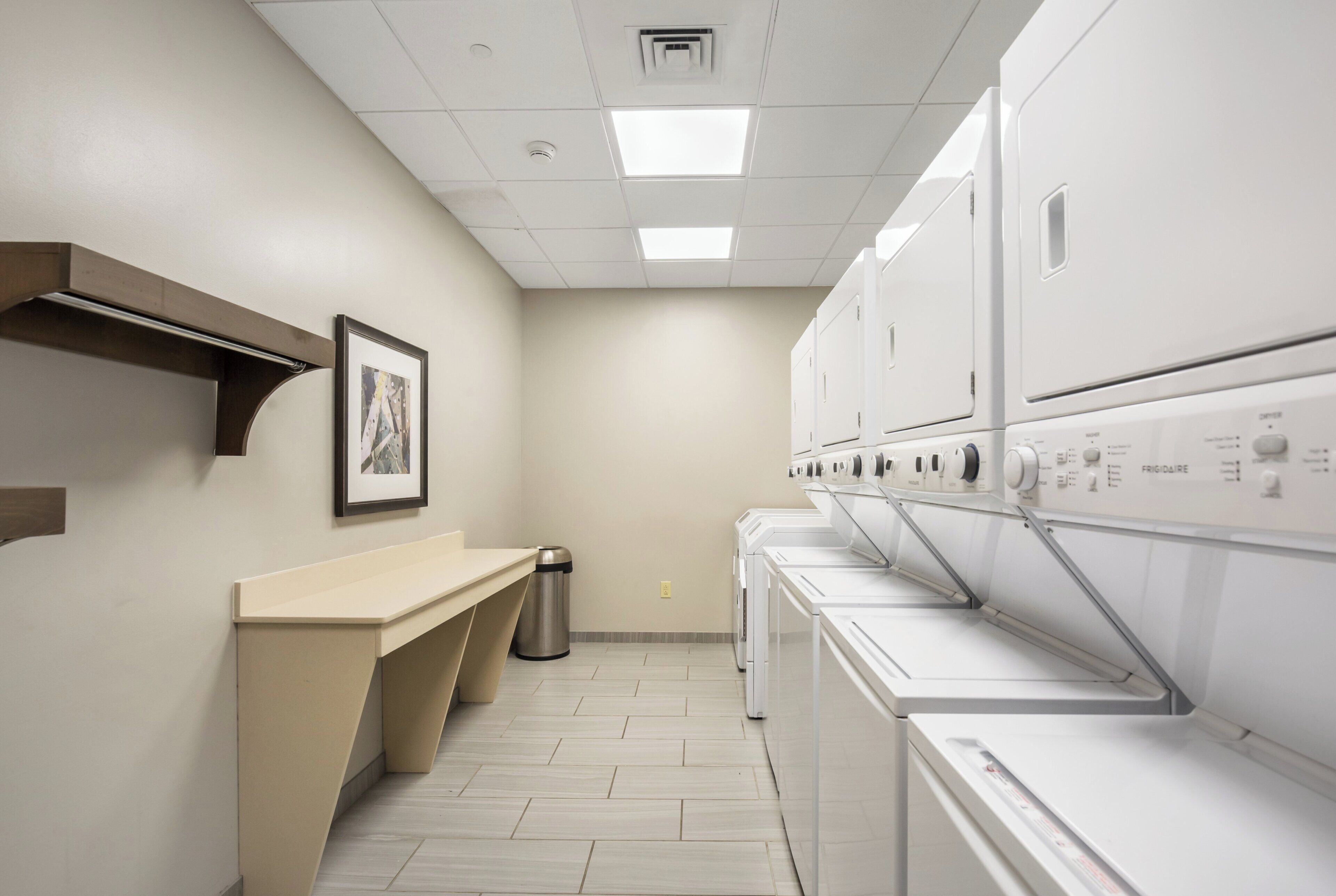 A laundry room with a row of white washers and dryers, a trash can, and a framed picture on the wall.