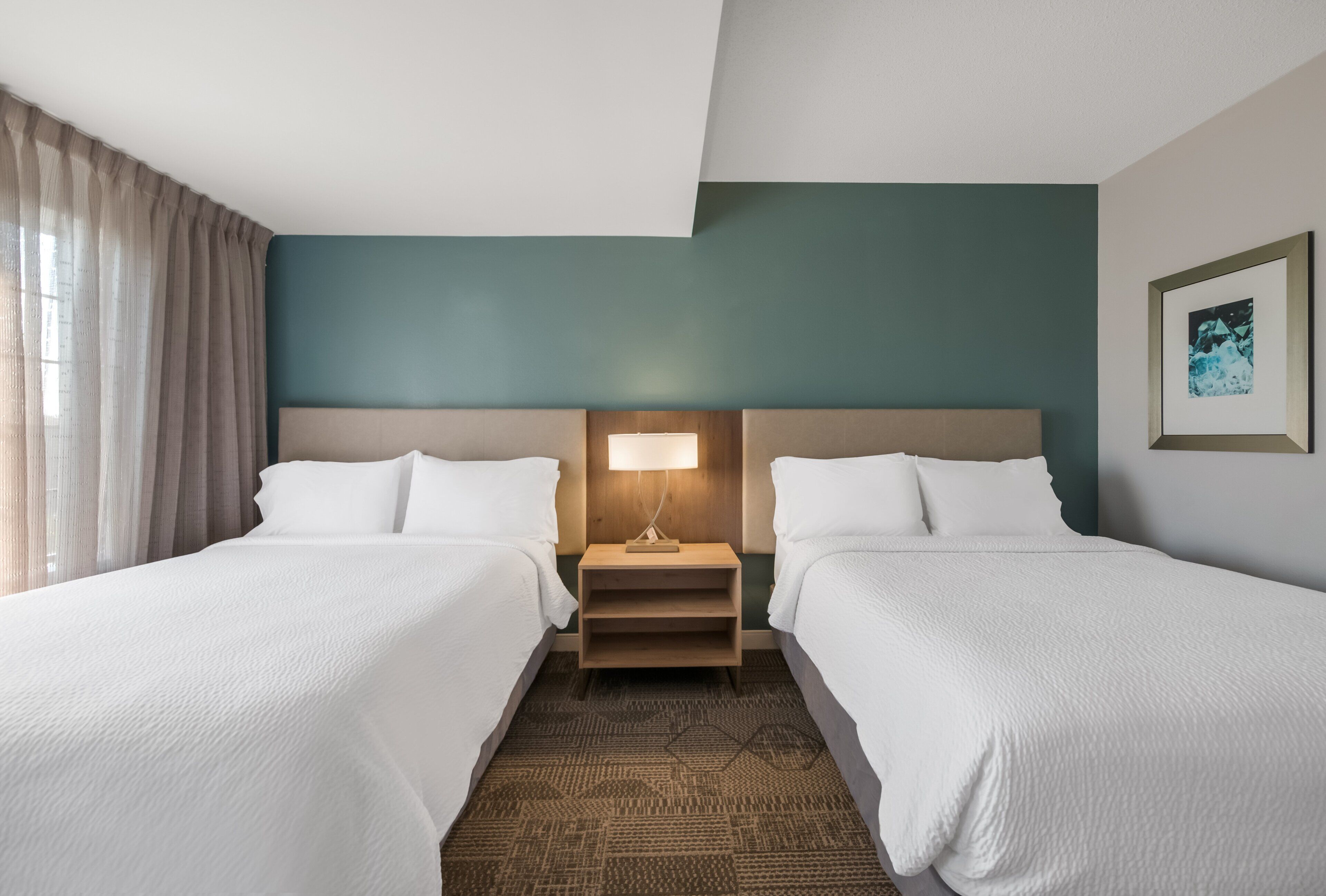 Une chambre d’hôtel avec deux lits, une table de chevet avec une lampe, un tableau encadré au mur et une fenêtre avec des rideaux.