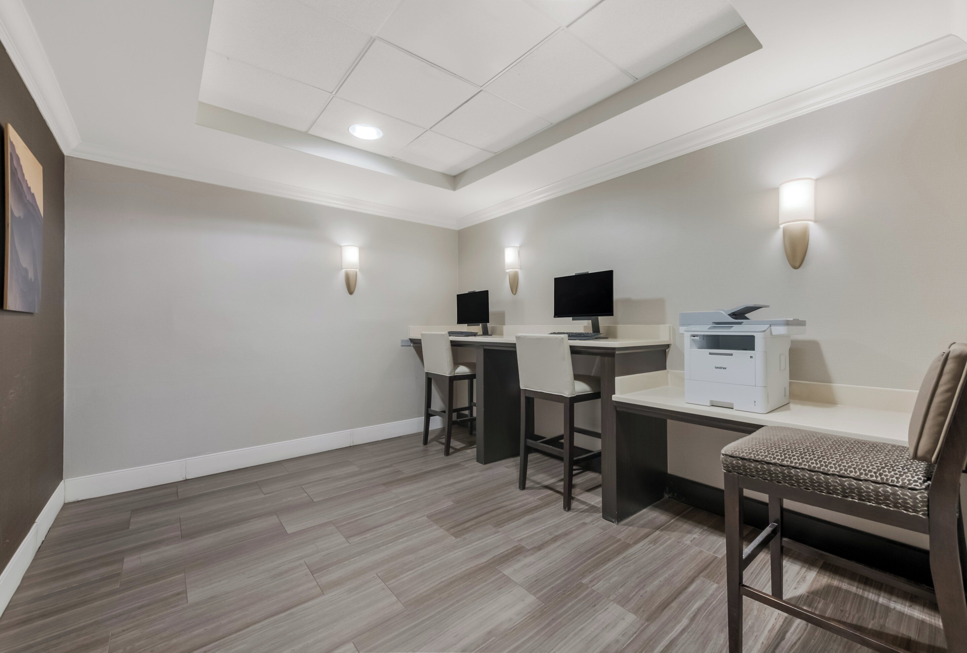 A hotel room with a desk, two chairs, a flat-screen TV, a printer, and a wall-mounted lamp.