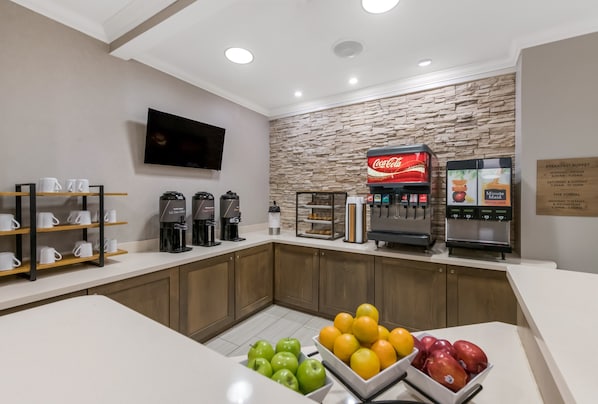A modern kitchen area with a stone wall, a flat-screen TV, a Coca-Cola refrigerator, a coffee machine, and a fruit bowl.