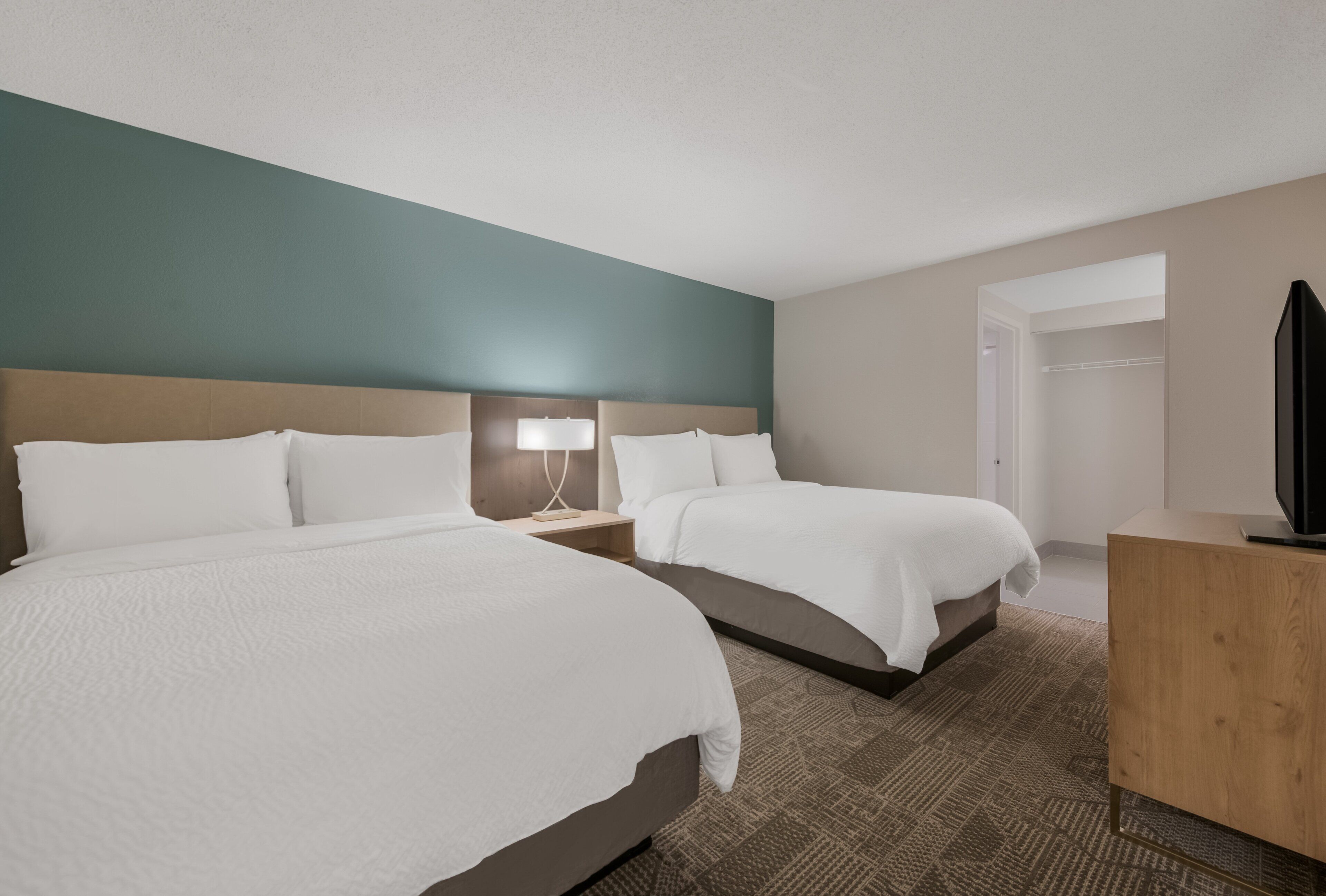 A hotel room with two beds, a bedside table with a lamp, a wooden dresser, and a television.