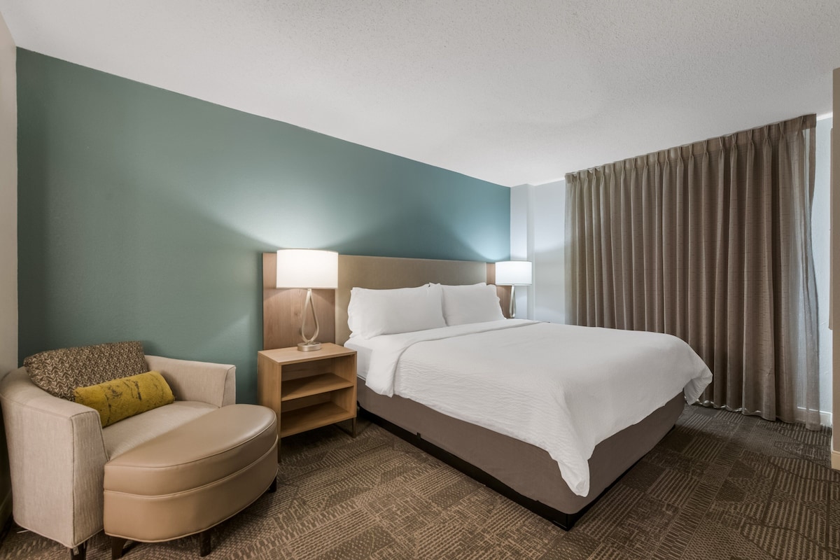 A hotel room with a bed, a chair, a nightstand, and a lamp.