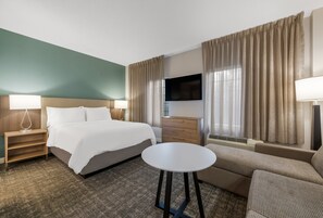 A hotel room with a bed, a sofa, a TV, a lamp, a bedside table, and a window with curtains. - Staybridge Suites Chesapeake by IHG (Chesapeake)