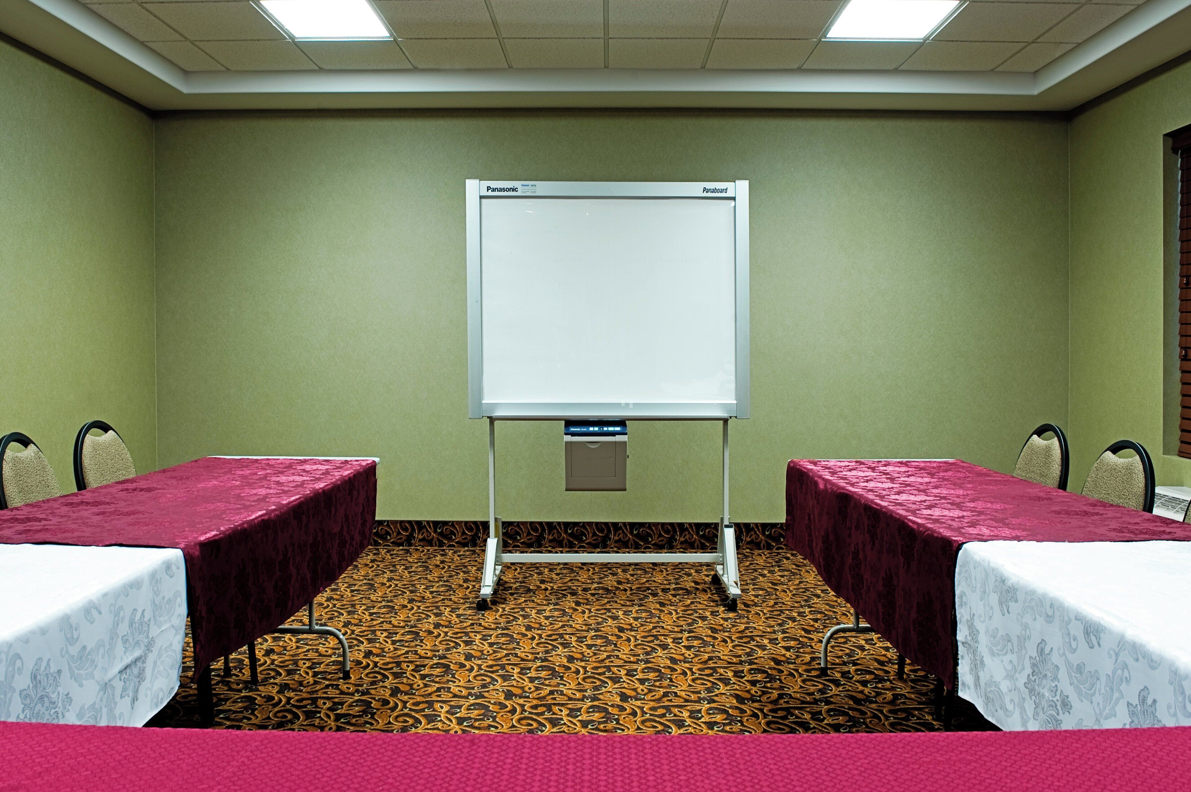 A conference room with a whiteboard, two tables with red tablecloths, patterned carpet, and green walls.