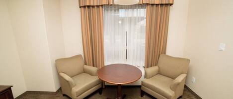 Habitación de hotel con dos sillones, una mesita redonda, ventana con cortinas e iluminación en el techo.