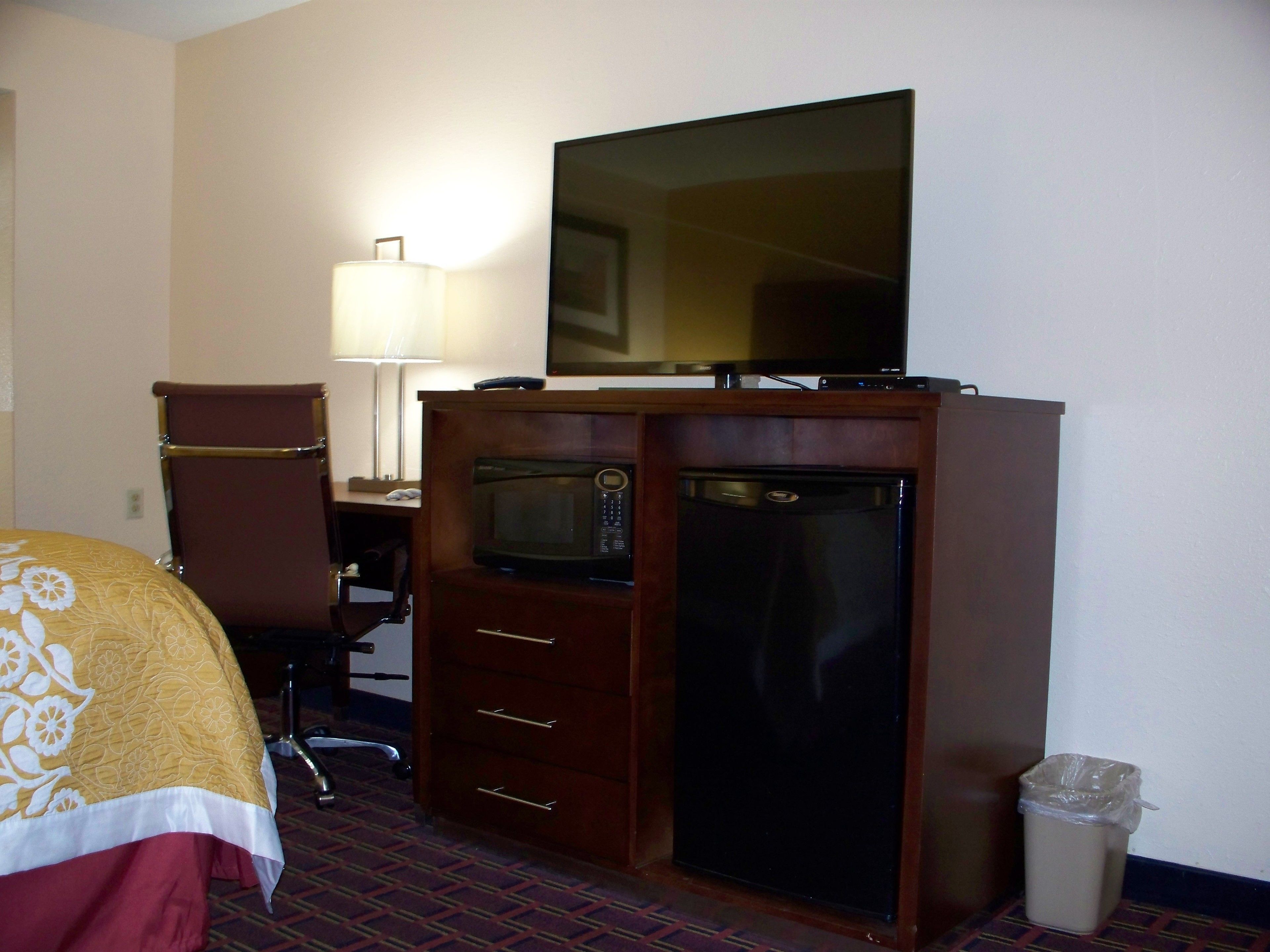 Una habitación de hotel con cama, escritorio con lámpara, televisor, microondas, refrigerador y basurero.
