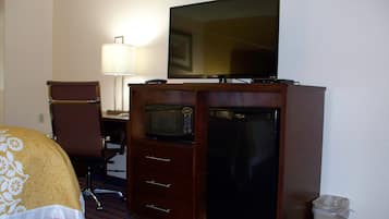 Una habitación de hotel con cama, escritorio con lámpara, televisor, microondas, refrigerador y basurero.