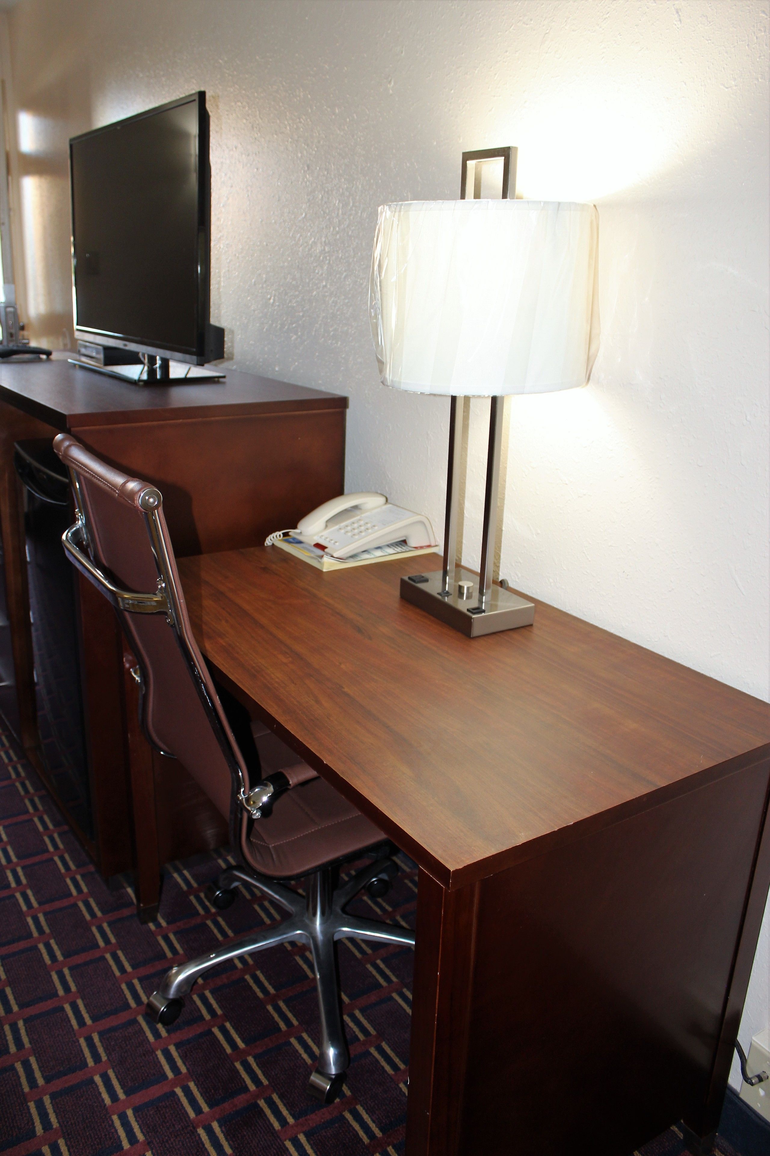 A hotel room with a wooden desk, a chair, a television, a lamp, and a telephone.