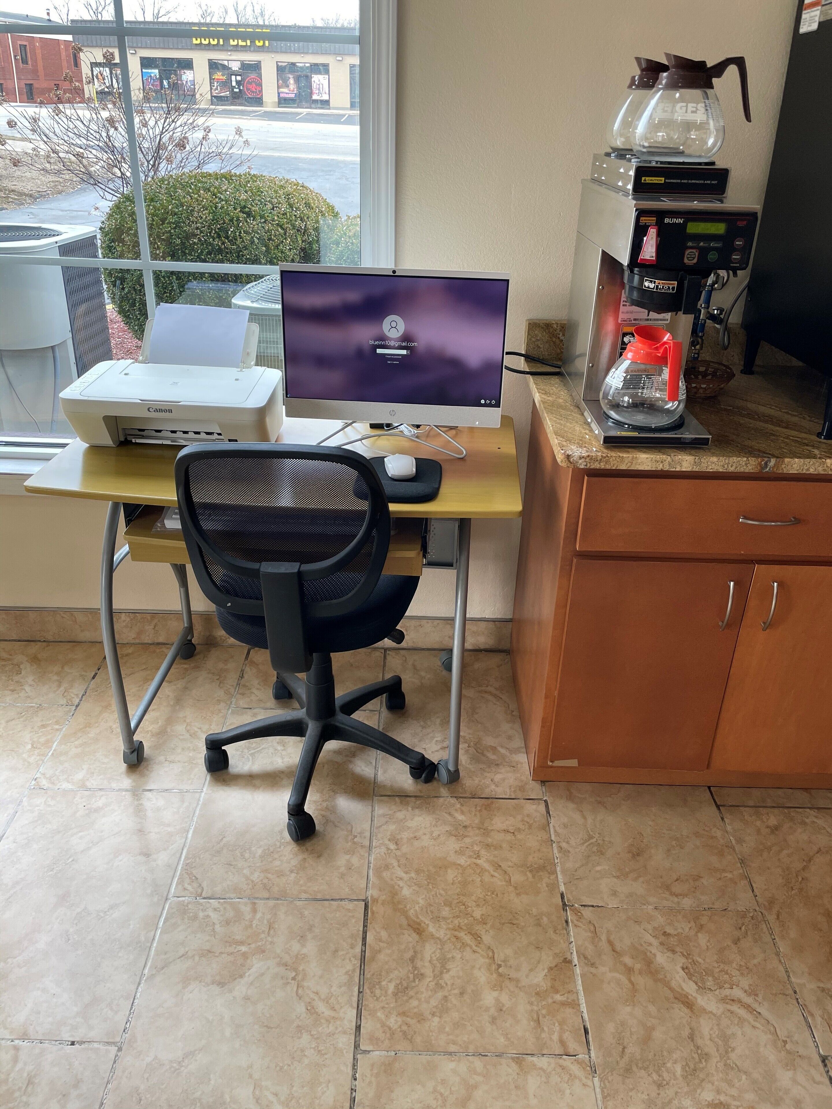 Um espaço de escritório em casa com um computador desktop, impressora, cafeteira e uma cadeira de escritório.
