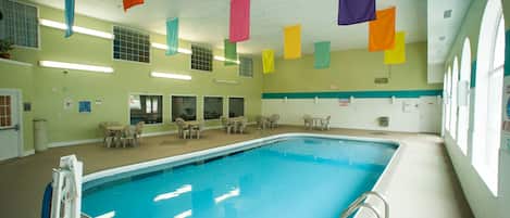 An indoor swimming pool with clear blue water, surrounded by lounge chairs and tables, and colorful flags hanging from the ceiling.