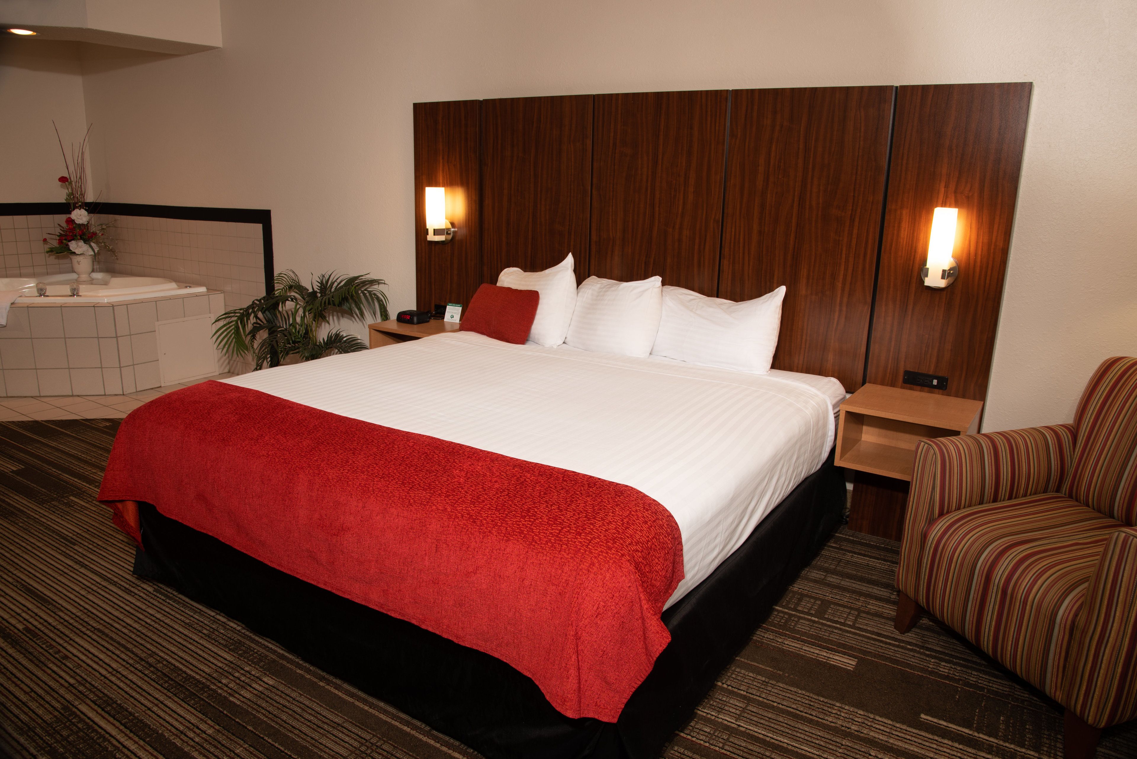 Une chambre d’hôtel avec un grand lit, une tête de lit en bois, deux tables de chevet, un plaid rouge, un fauteuil à rayures et une salle de bain avec un lavabo et un miroir.