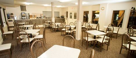 A spacious dining area with multiple tables and chairs, a kitchen in the background, and a clock on the wall.