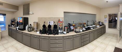Zona de desayuno autoservicio con máquinas de café, dispensador de fruta y diferentes recipientes de comida.