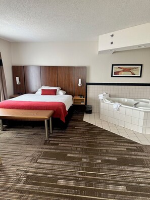 A hotel room with a bed, a bench, a small table, a nightstand, a wall-mounted light, a framed picture, a tiled wall, and a bathtub.