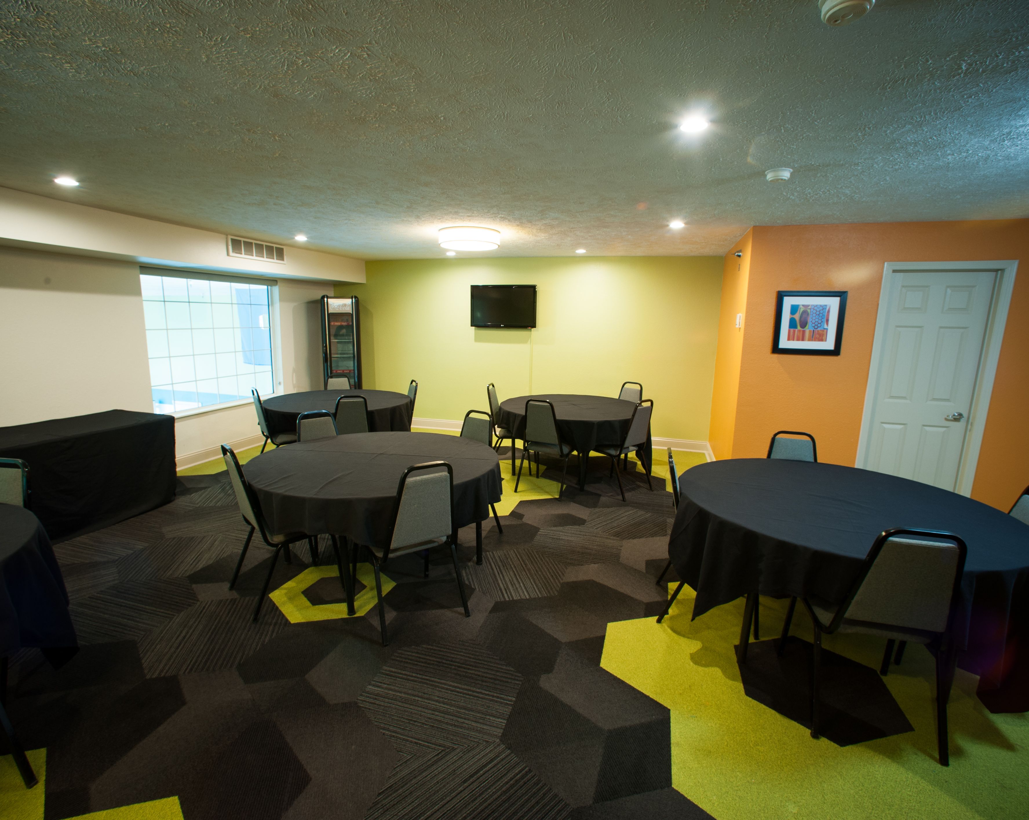 A conference room with multiple tables and chairs, a flat-screen TV on the wall, and a door in the background.