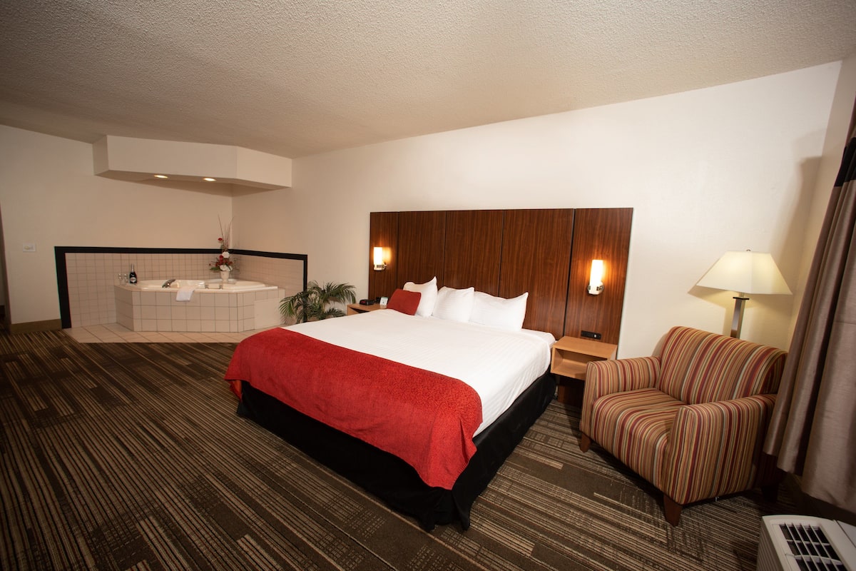 A hotel room with a large bed, a sitting area with a chair, a small table, a nightstand, a wall-mounted TV, and a bathroom with a bathtub and sink.