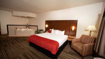 A hotel room with a large bed, a sitting area with a chair, a small table, a nightstand, a wall-mounted TV, and a bathroom with a bathtub and sink.
