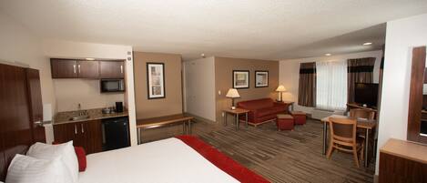 A hotel room with a bed, a desk, a chair, a sofa, a TV, a microwave, and a small kitchenette.