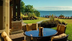 Une terrasse extérieure dotée d’une table ronde en verre et de chaises en osier, offrant une vue sur un jardin aménagé et l’océan.