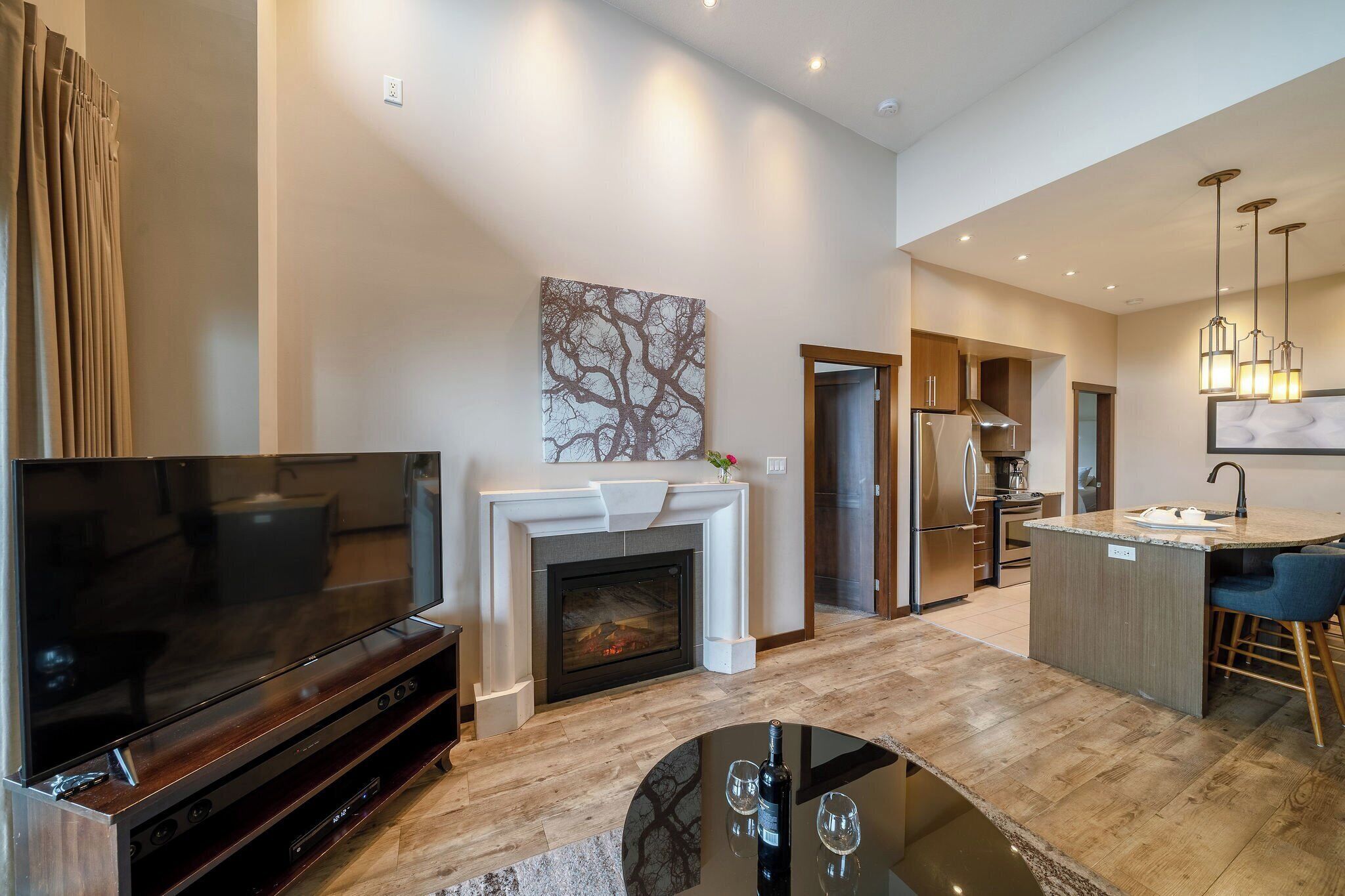 A modern living room with a flat-screen TV, a fireplace, and a dining area with a kitchen in the background.