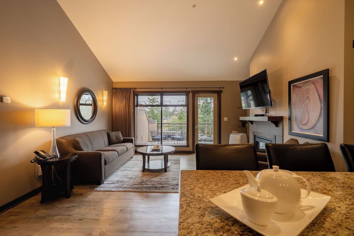 A modern hotel room with a sofa, a coffee table, a dining table with a teapot and cup, a TV, and a large window with a view of the outdoors.