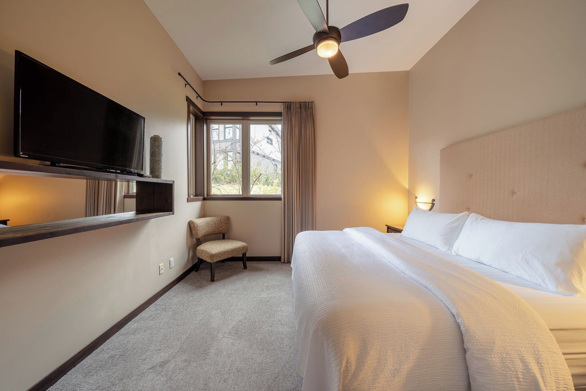 A hotel room with a bed, a television on a shelf, a chair, a window with curtains, and a ceiling fan.