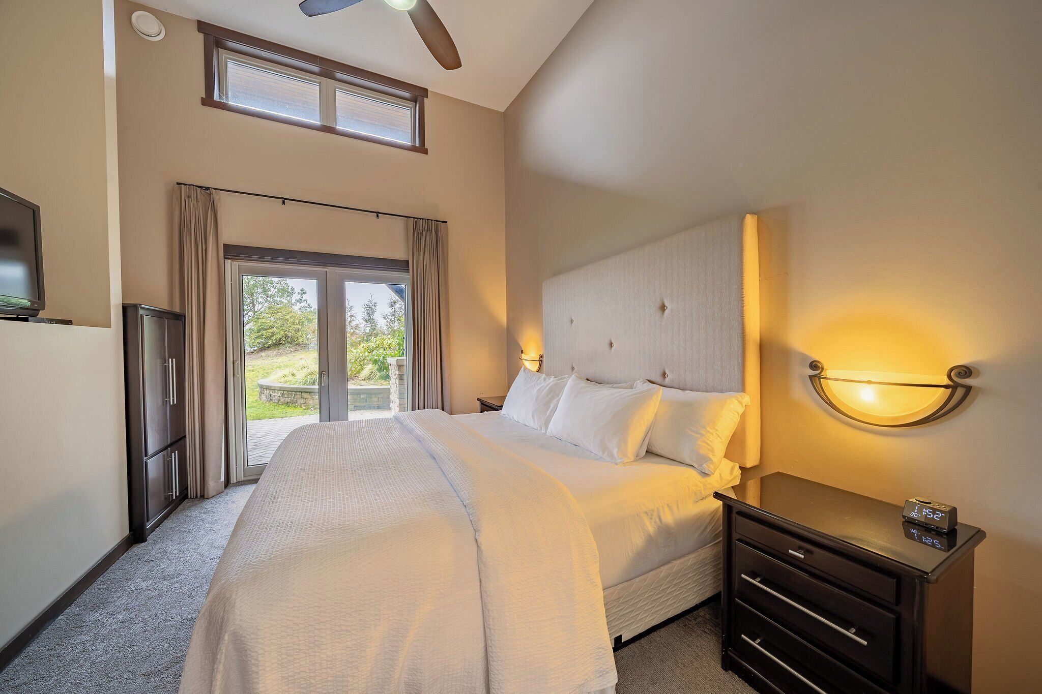 A hotel room with a large bed, a nightstand with a clock, a ceiling fan, and a view of the outdoors through a sliding door.
