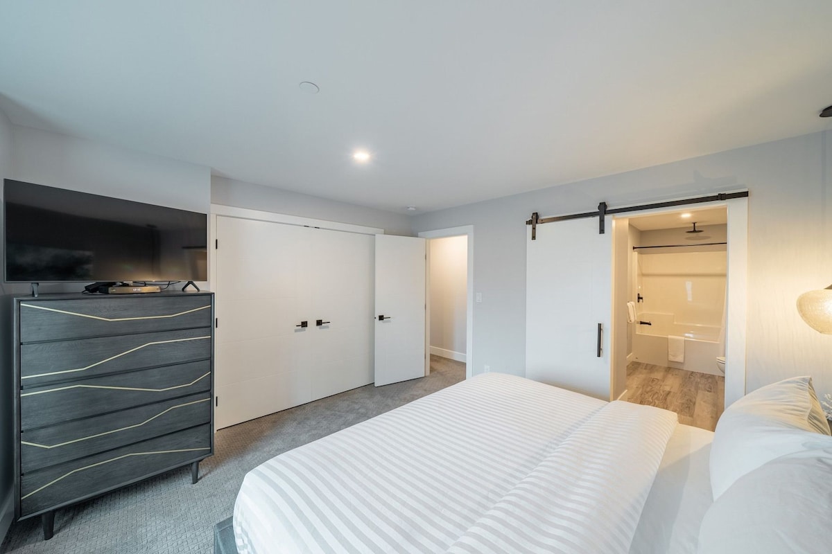 Un dormitorio moderno con cama, cómoda, televisor y puerta corrediza hacia un baño.