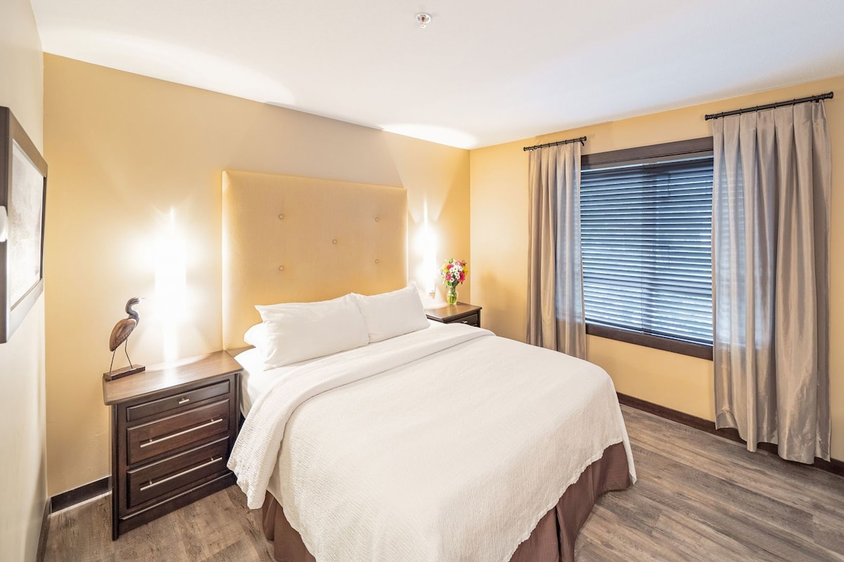 Habitación de hotel con cama, mesita de noche, lámpara y ventana con cortinas.