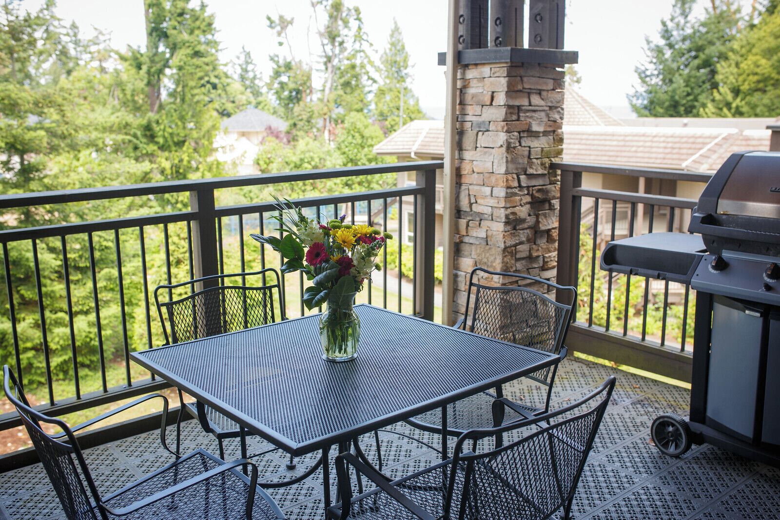 A balcony with a metal table and chairs, a vase of flowers, a stone pillar, and a grill.