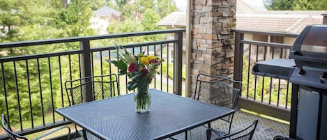 A balcony with a metal table and chairs, a vase of flowers, a stone pillar, and a grill.