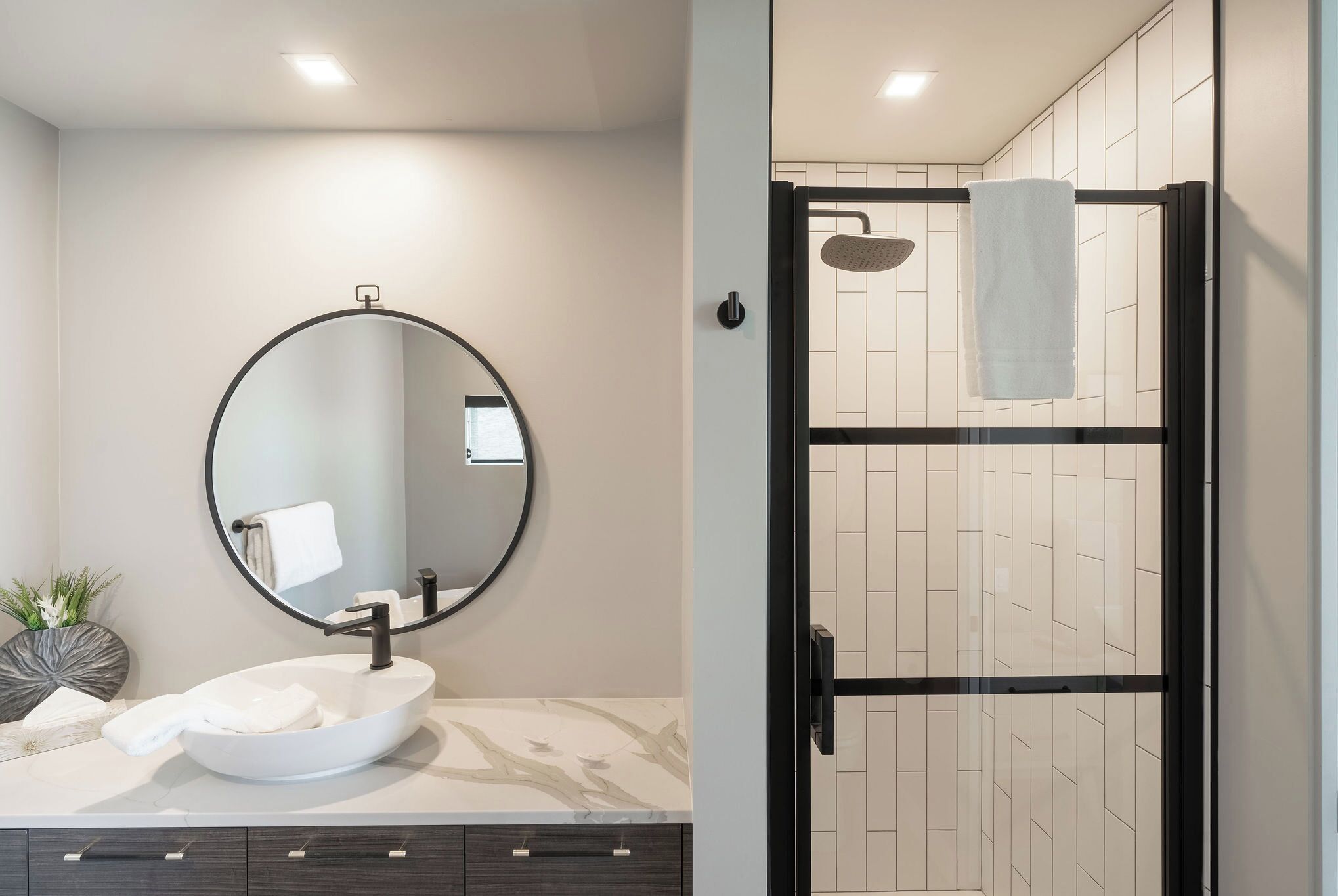 A modern bathroom with a round mirror, a white sink, dark wood cabinets, and a glass-enclosed shower area.