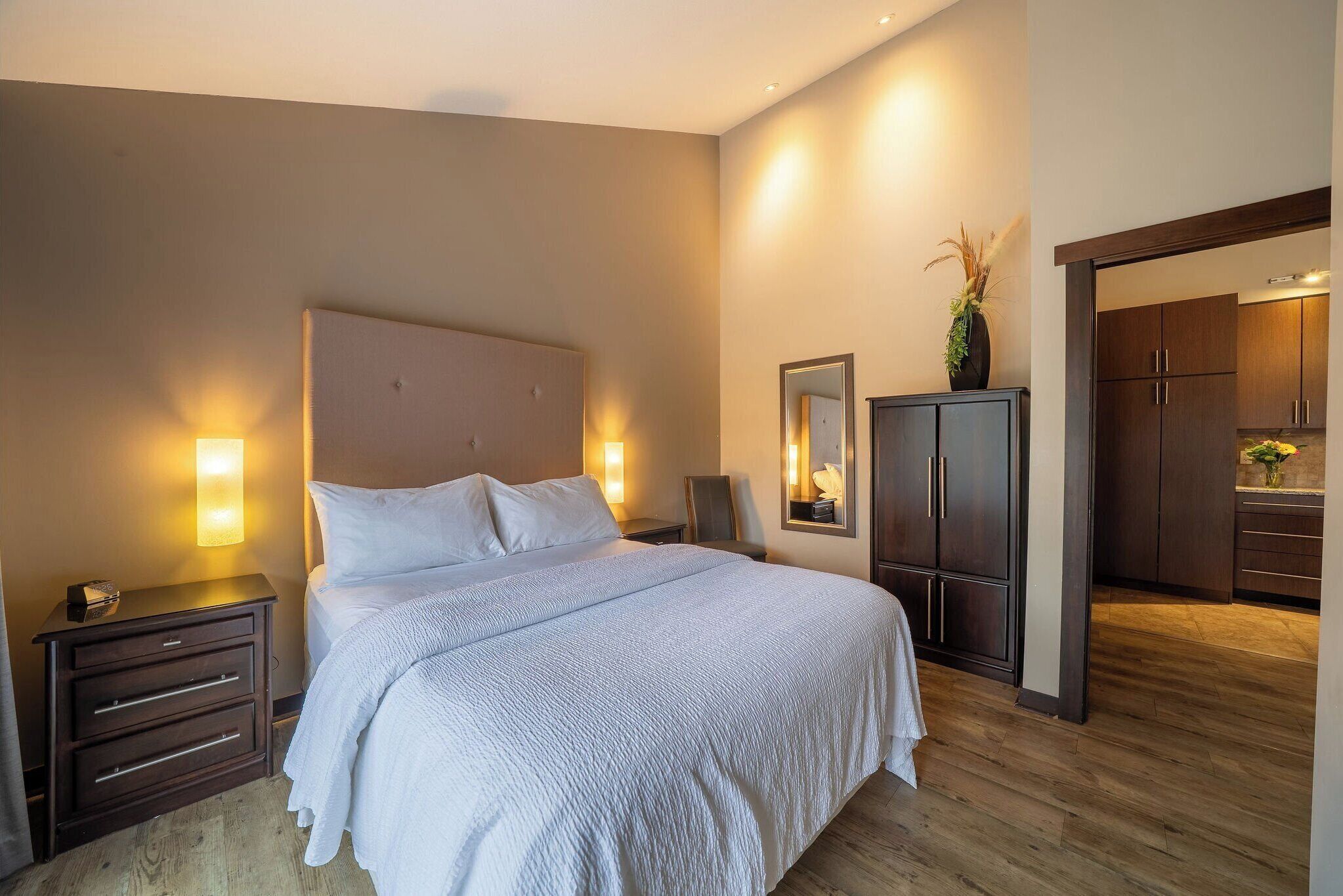 A hotel room with a large bed, wooden floor, a nightstand with a lamp, a wardrobe, and a mirror.